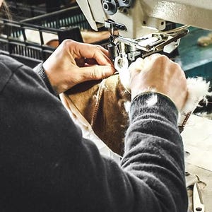 K&ouml;nnte beinhalten: Eine Person in einem schwarzen Langarmshirt bedient eine wei&szlig;e industrielle N&auml;hmaschine, um einen braunen Lederartikel mit wei&szlig;em Lammfellfutter zu n&auml;hen. Die Maschine tr&auml;gt das Etikett Lanmax LM9910-1T und einen runden Warnaufkleber.