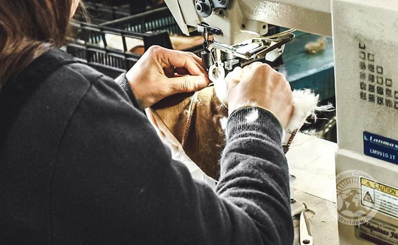 K&ouml;nnte beinhalten: Eine Person in einem schwarzen Langarmshirt bedient eine wei&szlig;e industrielle N&auml;hmaschine, um einen braunen Lederartikel mit wei&szlig;em Lammfellfutter zu n&auml;hen. Die Maschine tr&auml;gt das Etikett Lanmax LM9910-1T und einen runden Warnaufkleber.