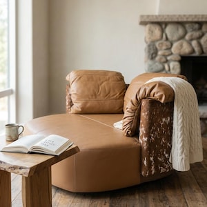 May include: A round, tan leather sofa with two large cushions. A white cable-knit blanket and a cowhide patterned accent are draped over the back. A small wooden side table holds an open book and a mug. The sofa sits on a hardwood floor.