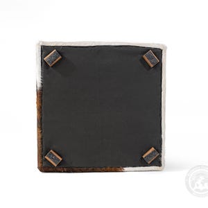May include: Square cowhide ottoman with a black underside and four wooden corner feet. The ottoman features brown and white fur on the sides and top edges. The underside is a solid black fabric.