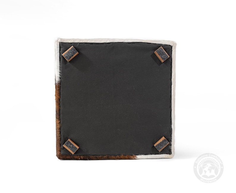 May include: Square cowhide ottoman with a black underside and four wooden corner feet. The ottoman features brown and white fur on the sides and top edges. The underside is a solid black fabric.