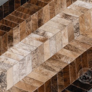 May include: A cowhide rug with a geometric pattern. The rug features a patchwork design with rectangular pieces in shades of brown, tan, and cream. The pieces are arranged in a chevron pattern, creating a visually interesting texture and design.