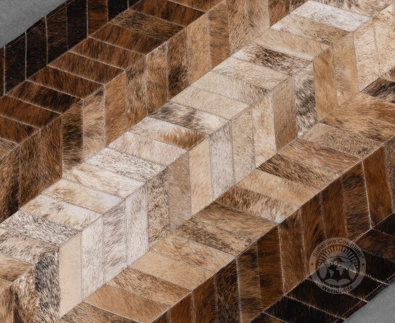 May include: A cowhide rug with a geometric pattern. The rug features a patchwork design with rectangular pieces in shades of brown, tan, and cream. The pieces are arranged in a chevron pattern, creating a visually interesting texture and design.