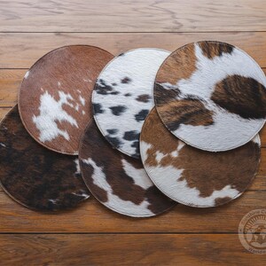 May include: A collection of six circular coasters featuring various cowhide patterns in shades of brown, white, and black. Each coaster has a stitched edge and a textured fur surface, displaying unique spotted and mottled animal print designs.