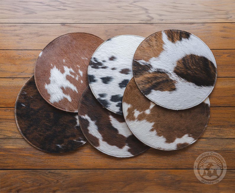 May include: A collection of six circular coasters featuring various cowhide patterns in shades of brown, white, and black. Each coaster has a stitched edge and a textured fur surface, displaying unique spotted and mottled animal print designs.