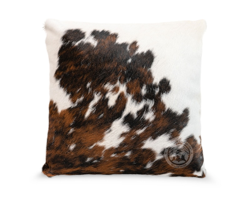 May include: A square throw pillow featuring a natural cowhide pattern with patches of white, dark brown, and black fur. The front surface displays a circular logo stamp in the bottom right corner with the text Cowhide Worldwide.