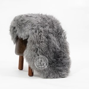 Puede incluir: Una alfombra de piel de oveja gris sobre un peque&ntilde;o taburete de cuero marr&oacute;n con patas de madera. La alfombra tiene una textura gruesa y peluda y un aspecto natural. El taburete es de dise&ntilde;o sencillo, con asiento marr&oacute;n oscuro y tres patas.