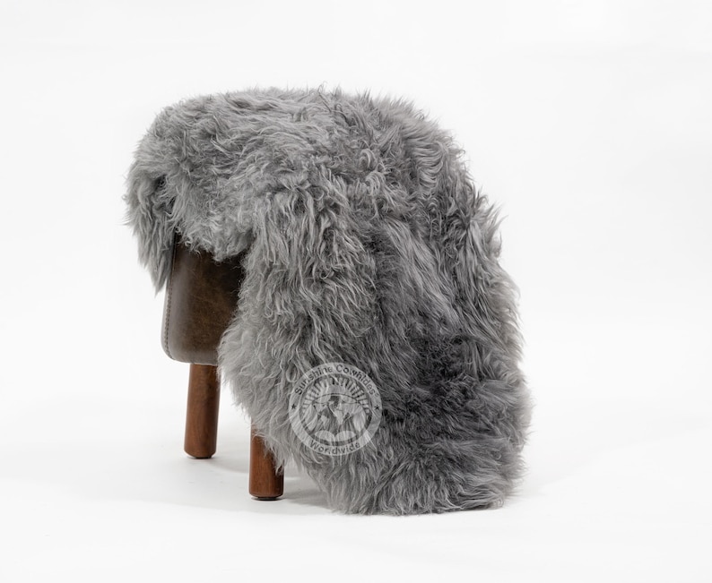Puede incluir: Una alfombra de piel de oveja gris sobre un peque&ntilde;o taburete de cuero marr&oacute;n con patas de madera. La alfombra tiene una textura gruesa y peluda y un aspecto natural. El taburete es de dise&ntilde;o sencillo, con asiento marr&oacute;n oscuro y tres patas.