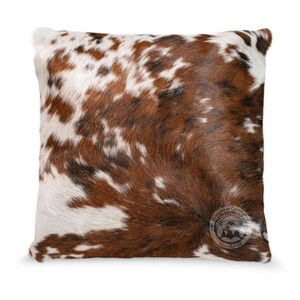 May include: A square throw pillow featuring a natural cowhide pattern with a mix of brown, white, and black patches. The surface has a textured hair-on-hide appearance. A circular logo with the text Sunshine Cowhide Worldwide is visible.