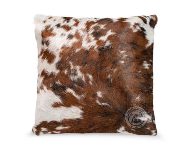 May include: A square throw pillow featuring a natural cowhide pattern with a mix of brown, white, and black patches. The surface has a textured hair-on-hide appearance. A circular logo with the text Sunshine Cowhide Worldwide is visible.