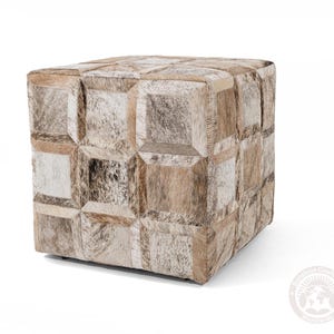 May include: A square ottoman with a patchwork design made from cowhide. The ottoman features a variety of neutral tones, including beige, brown, and white, arranged in a geometric pattern. The piece is a cube shape and appears to be sturdy.