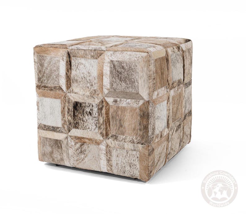 May include: A square ottoman with a patchwork design made from cowhide. The ottoman features a variety of neutral tones, including beige, brown, and white, arranged in a geometric pattern. The piece is a cube shape and appears to be sturdy.