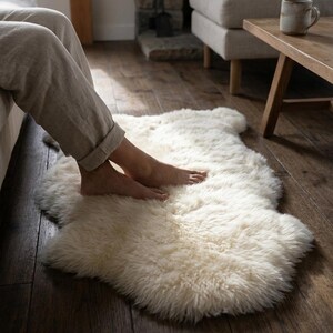 Puede incluir: Una alfombra de piel de oveja color crema con una textura suave y esponjosa. La alfombra est&aacute; colocada sobre un suelo de madera oscura, con pies descalzos descansando sobre ella. La alfombra a&ntilde;ade un elemento acogedor a la habitaci&oacute;n.