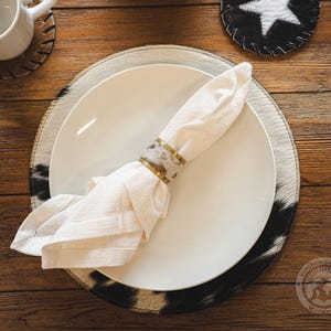 May include: A white cloth napkin folded and secured with a cylindrical napkin ring featuring a metallic gold rim and a central band of brown and white cowhide pattern, resting on a white ceramic plate atop a round cowhide placemat.