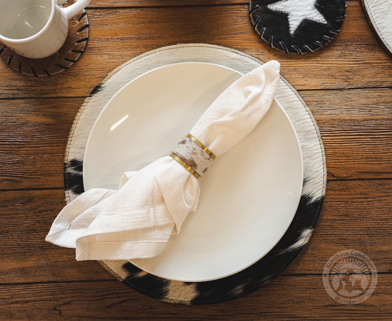 May include: A white cloth napkin folded and secured with a cylindrical napkin ring featuring a metallic gold rim and a central band of brown and white cowhide pattern, resting on a white ceramic plate atop a round cowhide placemat.