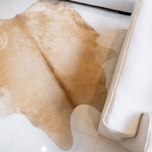 May include: A light brown cowhide rug with a natural, irregular shape. The rug is laid out on a white tiled floor and partially covers a white sofa.