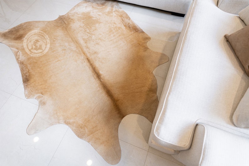 May include: A light brown cowhide rug with a natural, irregular shape. The rug is laid out on a white tiled floor and partially covers a white sofa.