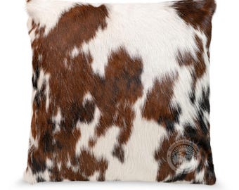 May include: A square throw pillow featuring a natural cowhide pattern with irregular patches of brown, white, and black fur. The bottom right corner displays a circular logo with the text Sunshine Cowhides printed in a dark font.