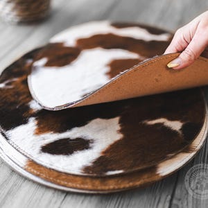 May include: A stack of circular placemats featuring a brown and white cowhide pattern. The top placemat is lifted to reveal a smooth, tan-colored underside. Each placemat has a stitched edge, and the set is arranged on a wooden surface.