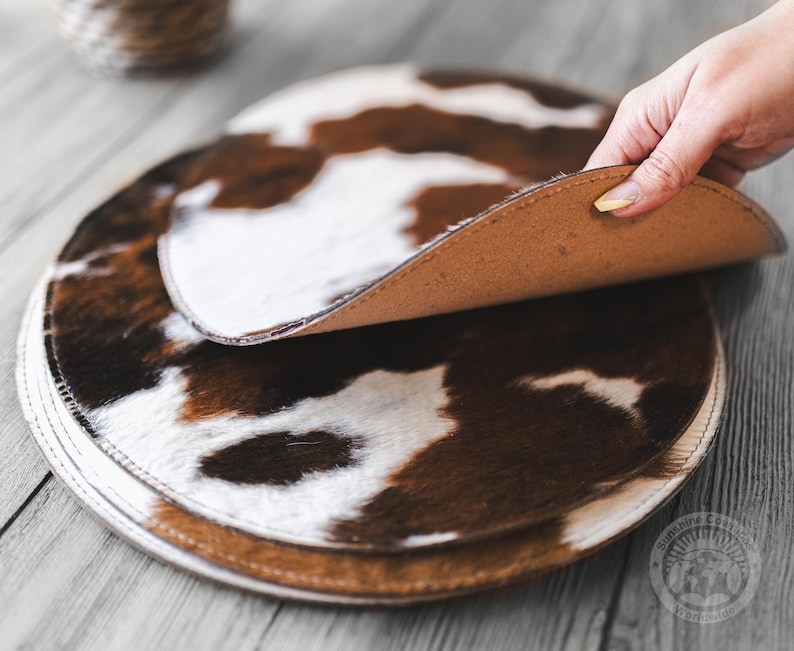 May include: A stack of circular placemats featuring a brown and white cowhide pattern. The top placemat is lifted to reveal a smooth, tan-colored underside. Each placemat has a stitched edge, and the set is arranged on a wooden surface.