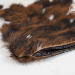 May include: A rectangular pouch featuring a textured brown, black, and white animal fur exterior. The pouch includes a dark brown zipper closure along the top edge. A circular logo with the text Sunshine Country Worldwide is visible in the corner.