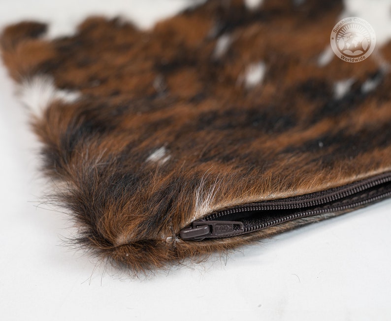 May include: A rectangular pouch featuring a textured brown, black, and white animal fur exterior. The pouch includes a dark brown zipper closure along the top edge. A circular logo with the text Sunshine Country Worldwide is visible in the corner.
