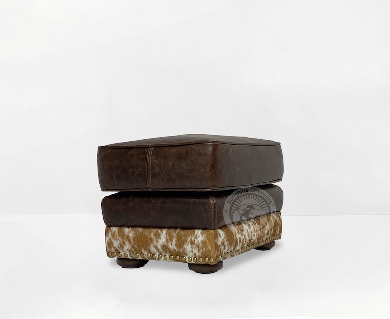 May include: A brown leather ottoman with a cowhide pattern on the bottom. The ottoman has a square shape and is decorated with gold studs.
