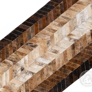 May include: A close-up of a rug with a herringbone pattern. The rug is made of cowhide in shades of brown, tan, and cream. The pattern is made up of rectangular pieces of hide, creating a textured surface. The rug has a natural, rustic look.