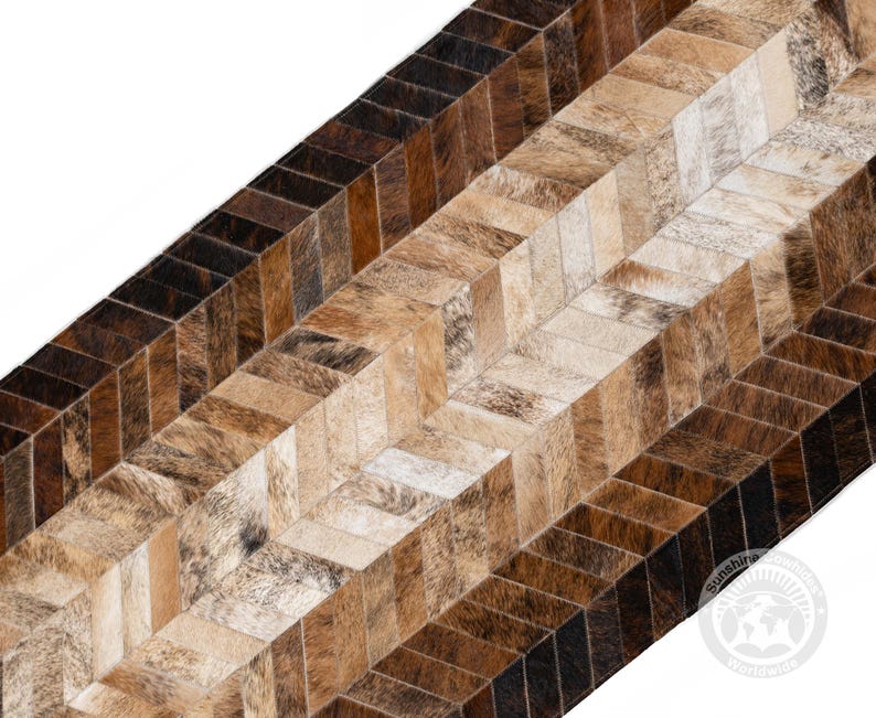 May include: A close-up of a rug with a herringbone pattern. The rug is made of cowhide in shades of brown, tan, and cream. The pattern is made up of rectangular pieces of hide, creating a textured surface. The rug has a natural, rustic look.