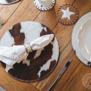 May include: A round placemat featuring a brown and white cowhide pattern, paired with a white cloth napkin held by a metallic napkin ring. A circular coaster with a white star on a brown cowhide background is positioned nearby on the wooden table.