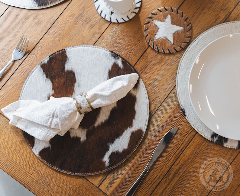 May include: A round placemat featuring a brown and white cowhide pattern, paired with a white cloth napkin held by a metallic napkin ring. A circular coaster with a white star on a brown cowhide background is positioned nearby on the wooden table.
