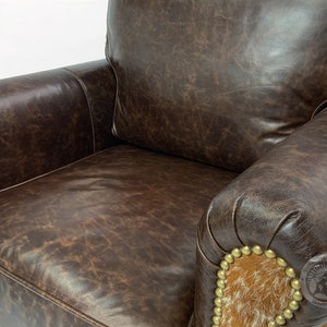 May include: A brown leather armchair with a distressed finish. The chair has a high back and rolled arms. The arms are accented with a brown and white cowhide patch and gold-toned studs.