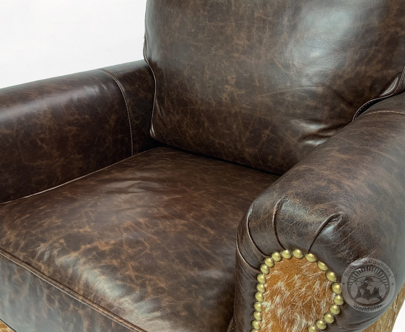May include: A brown leather armchair with a distressed finish. The chair has a high back and rolled arms. The arms are accented with a brown and white cowhide patch and gold-toned studs.