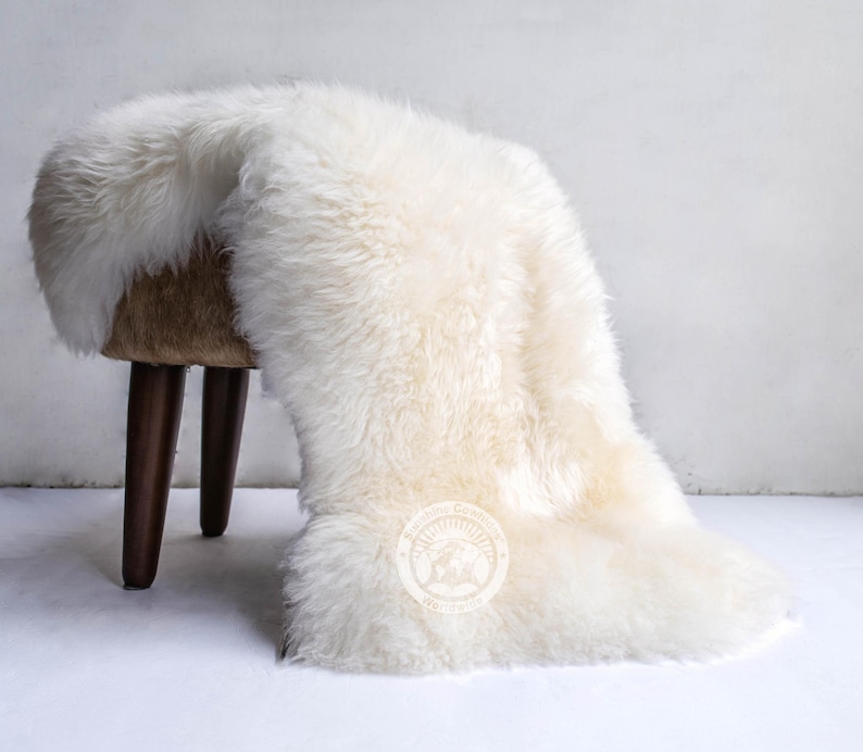 Puede incluir: Una alfombra de piel de oveja color crema est&aacute; sobre un peque&ntilde;o taburete redondo con patas marrones oscuras. La alfombra tiene fibras largas y esponjosas y una textura suave y acogedora. El taburete es de madera y tiene un acabado natural.