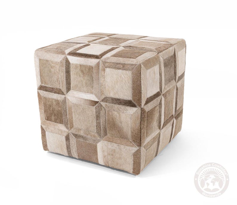 May include: A square ottoman with a patchwork design. The ottoman is covered in a mix of light and dark brown cowhide squares, creating a geometric pattern. The piece is a neutral color and would fit in many home decor styles.