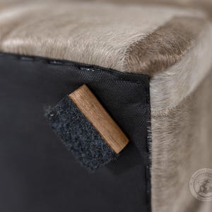 Pu&ograve; includere: Primo piano di un pouf in pelle di mucca di colore marrone chiaro con una base in tessuto nero e un blocco di legno attaccato alla parte inferiore. Il blocco di legno ha un cuscinetto in feltro nero sul fondo.