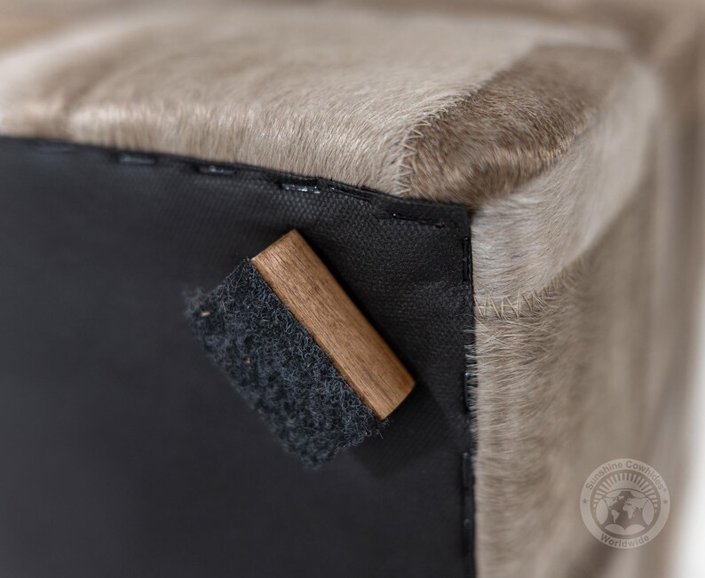 Pu&ograve; includere: Primo piano di un pouf in pelle di mucca di colore marrone chiaro con una base in tessuto nero e un blocco di legno attaccato alla parte inferiore. Il blocco di legno ha un cuscinetto in feltro nero sul fondo.
