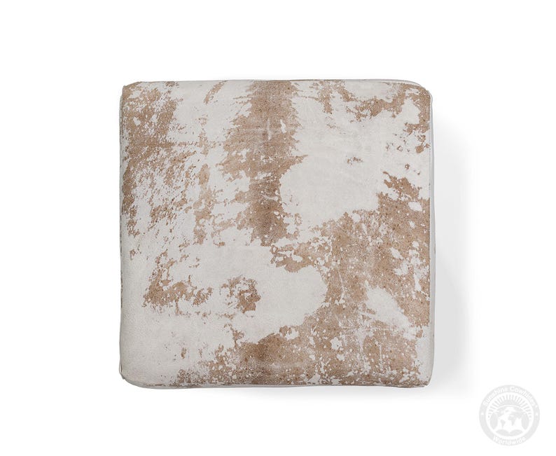 Puede incluir: Un coj&iacute;n de suelo cuadrado con un patr&oacute;n de piel de vaca natural con parches irregulares de pelo blanco y marr&oacute;n claro. El coj&iacute;n tiene una superficie superior plana y bordes definidos, mostrando una textura org&aacute;nica moteada en el panel.