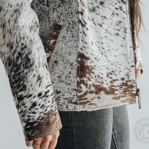 May include: A cowhide jacket with a zipper closure. The jacket features a pattern of white, brown, and black spots. The sleeve and pocket are visible. The jacket is a neutral color and can be worn with many outfits.