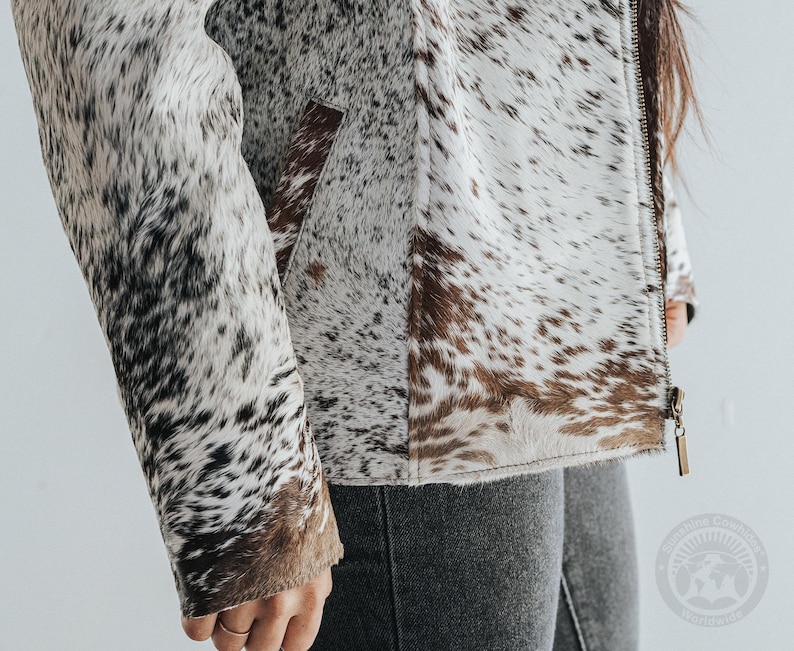 May include: A cowhide jacket with a zipper closure. The jacket features a pattern of white, brown, and black spots. The sleeve and pocket are visible. The jacket is a neutral color and can be worn with many outfits.