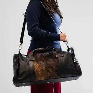May include: A black leather duffel bag featuring a central panel of brown and black animal hide with a textured fur finish. The bag includes dual top handles, a detachable shoulder strap with metal hardware, and silver-toned metal feet on the base.