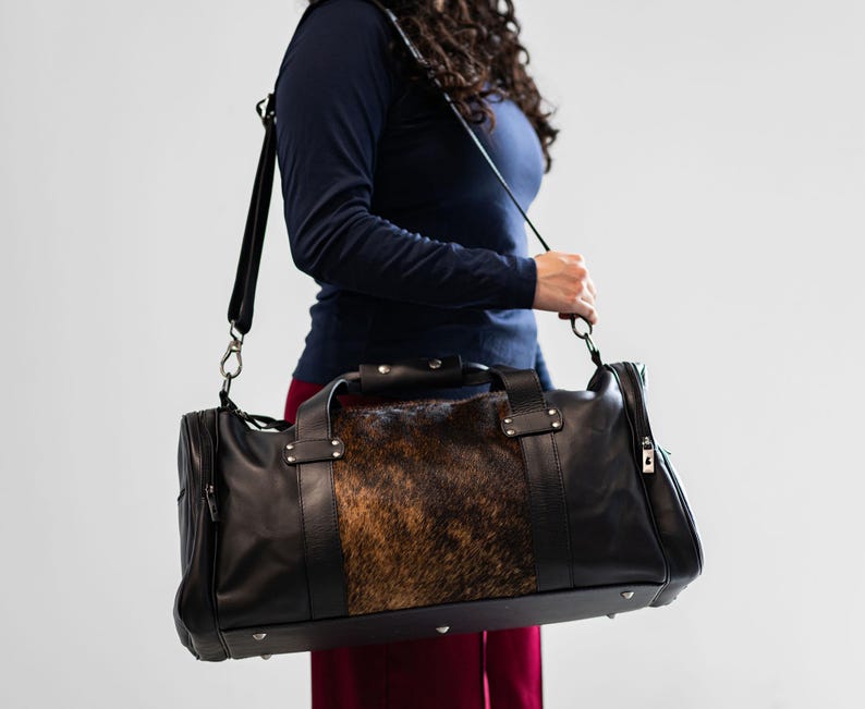 May include: A black leather duffel bag featuring a central panel of brown and black animal hide with a textured fur finish. The bag includes dual top handles, a detachable shoulder strap with metal hardware, and silver-toned metal feet on the base.
