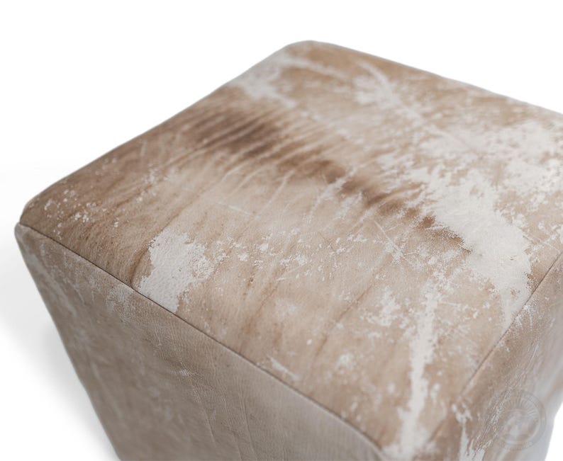 Puede incluir: Un puf cuadrado con un acabado desgastado en tonos marr&oacute;n claro y blanco. La superficie muestra una textura moteada y envejecida con patrones de grano visibles en la parte superior y los lados, creando una apariencia tonal en este mueble c&uacute;bico.