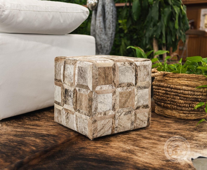 K&ouml;nnte beinhalten: Ein quadratischer Hocker mit einem Patchwork-Design aus braunem und wei&szlig;em Kuhfell. Der Hocker steht auf einem braunen Kuhfellteppich.