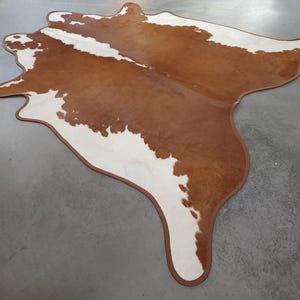 May include: A brown and white cowhide rug with a unique shape. The rug has a brown border and a natural pattern of brown and white patches. The rug is on a gray surface.
