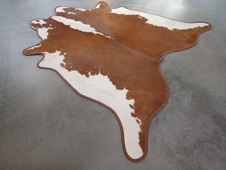 May include: A brown and white cowhide rug with a unique shape. The rug has a brown border and a natural pattern of brown and white patches. The rug is on a gray surface.