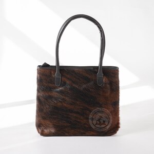 May include: A brown and black cowhide tote bag with black leather handles and a small leather tag that says "Genuine Cowhide Worldwide".