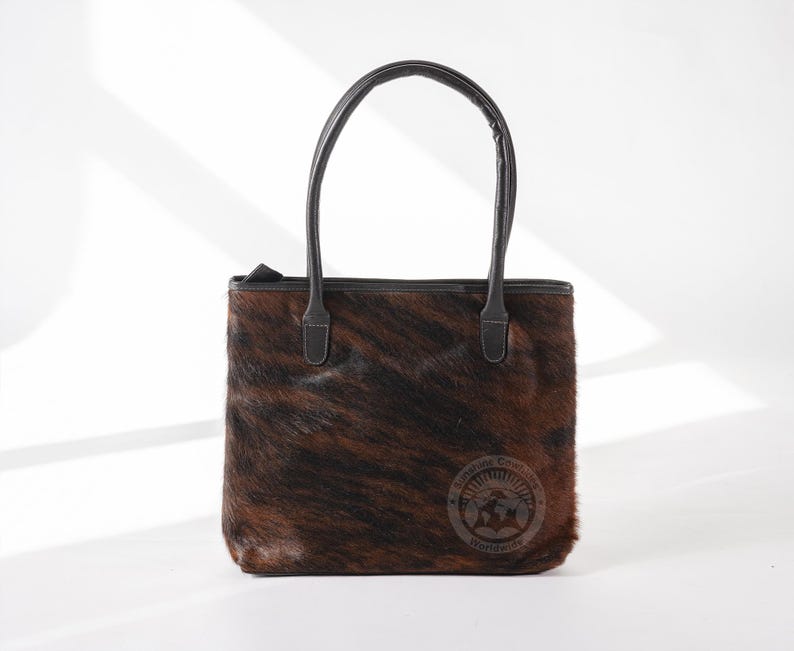 May include: A brown and black cowhide tote bag with black leather handles and a small leather tag that says "Genuine Cowhide Worldwide".
