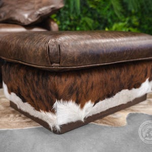May include: A brown leather ottoman with a cowhide pattern on the bottom. The ottoman is sitting on a light brown rug.