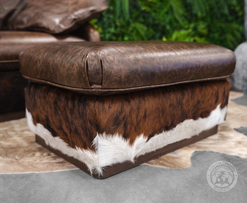 May include: A brown leather ottoman with a cowhide pattern on the bottom. The ottoman is sitting on a light brown rug.